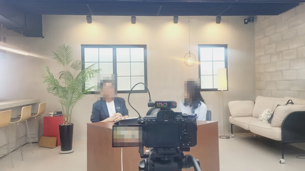 완성된 갈색 테이블에 두 사람이 마주 앉아 이야기하고 앞쪽에서 카메라가 촬영하는 현장, 인물 얼굴은 모자이크 처리